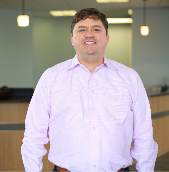 Professional man in lavender shirt smiling in modern office lobby setting.
