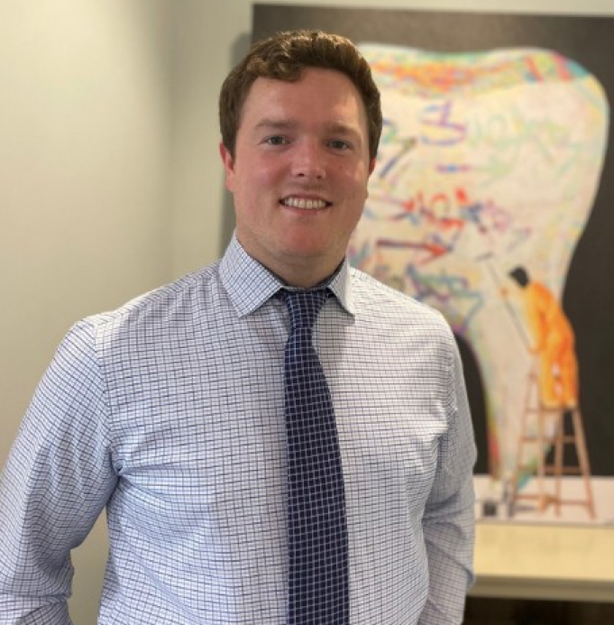 Professional man in business attire smiling in dental office with artwork on wall behind him.