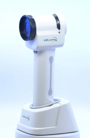 White handheld dental imaging device with blue lens mounted on charging base against white background.