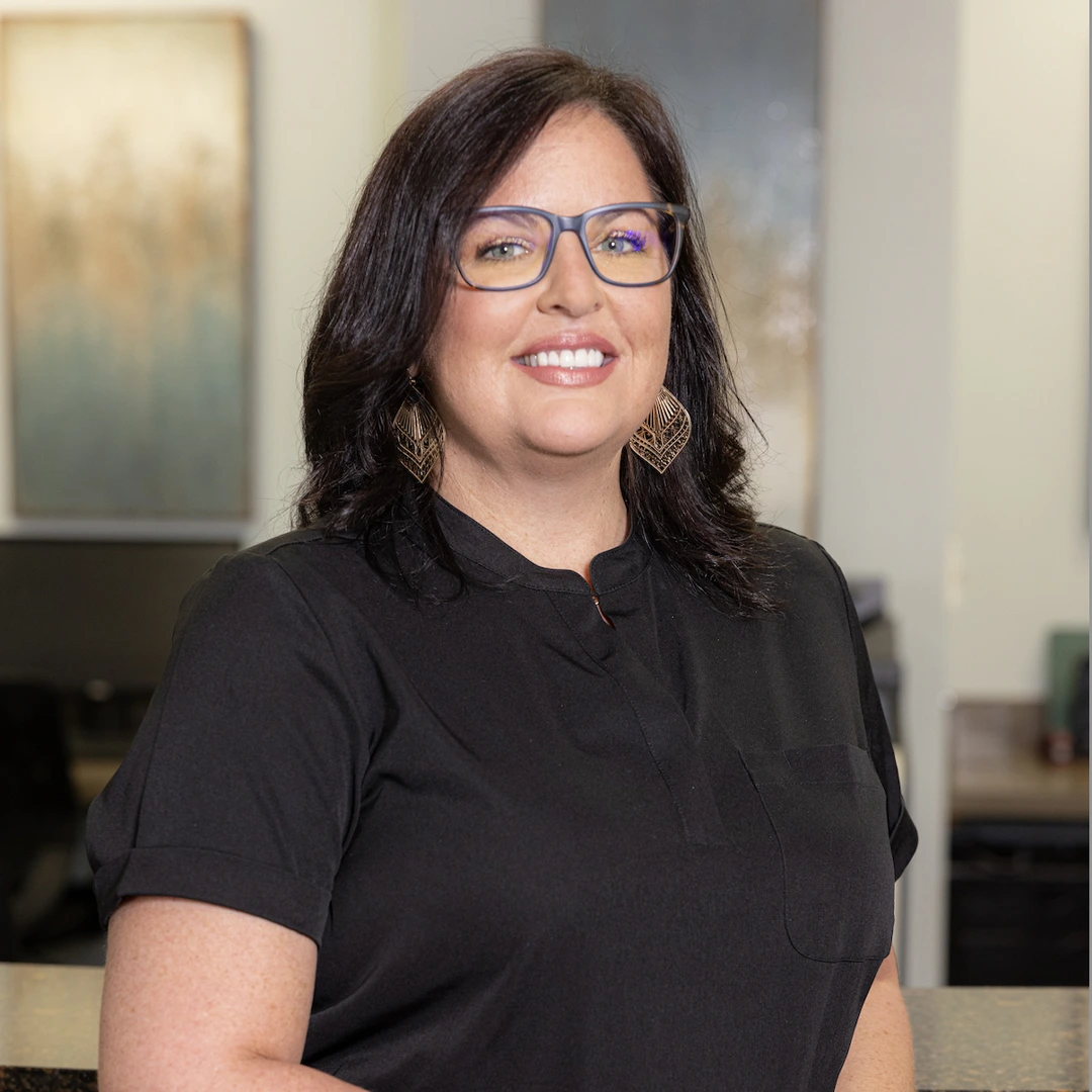 Woman wearing glasses and black polo shirt smiling in professional dental office setting.