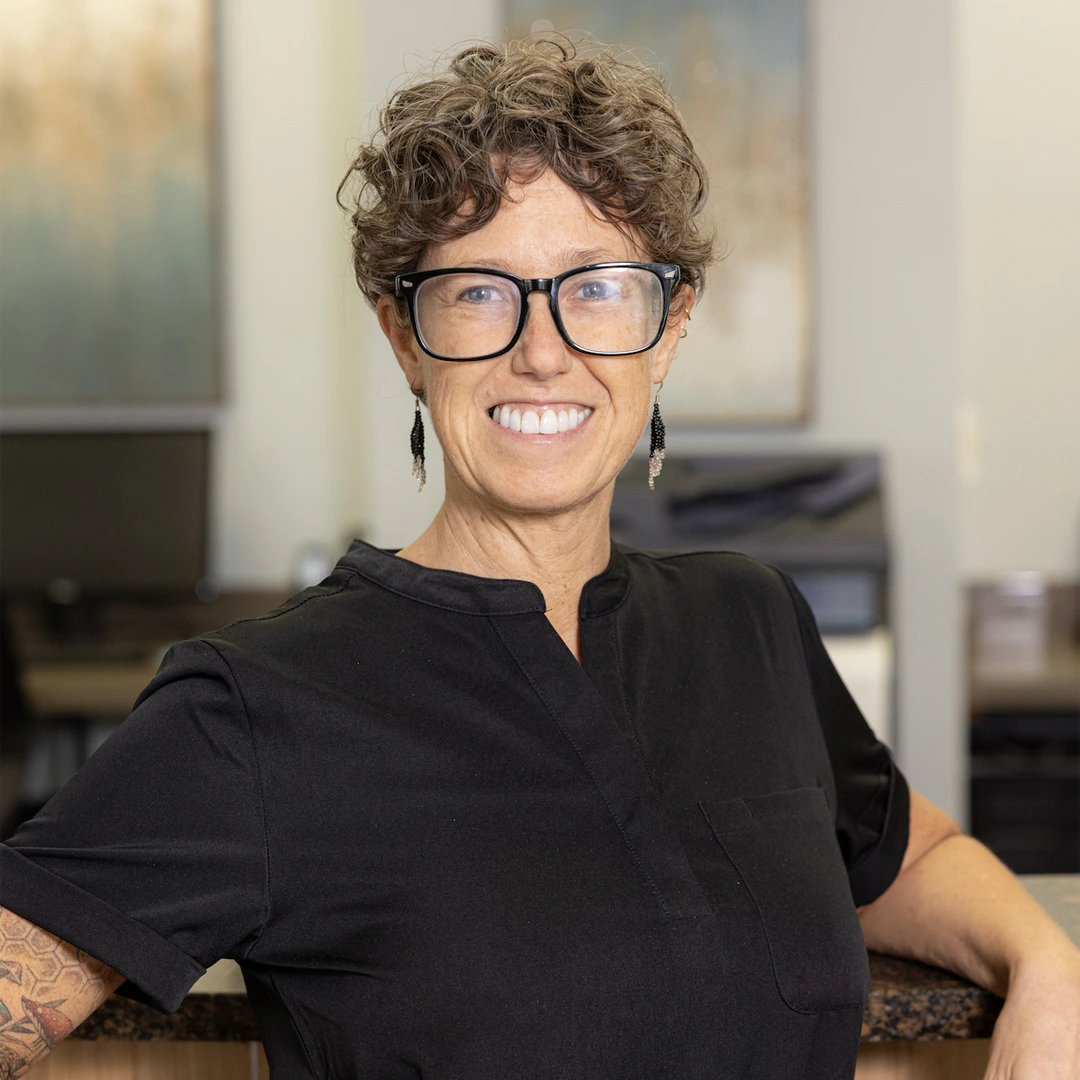 Professional woman with curly hair and glasses smiling in modern dental office setting.